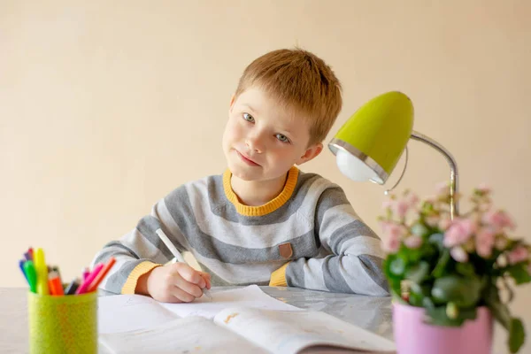 その少年は宿題をする。子供は遠隔で学んでいる。少年はテーブルに座って書き込みます。オンライン学習 — ストック写真