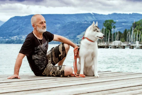 수염난 회색 머리의 남자와 그 의 개. 한 남자가 시베리아 허스키와 부두에 앉아 있습니다. 호수 근처에서 쉬 세요. 개 랑 산에 가는 여행 말이야. 빅토리 아호 아터 제. — 스톡 사진