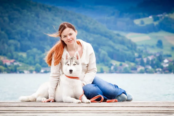 Una joven con cabello castaño y suéter blanco está sentada en el muelle del lago con el agua tranquila. Una perra hembra husky siberiana está acostada cerca de la chica. Las montañas de los Alpes al fondo . —  Fotos de Stock