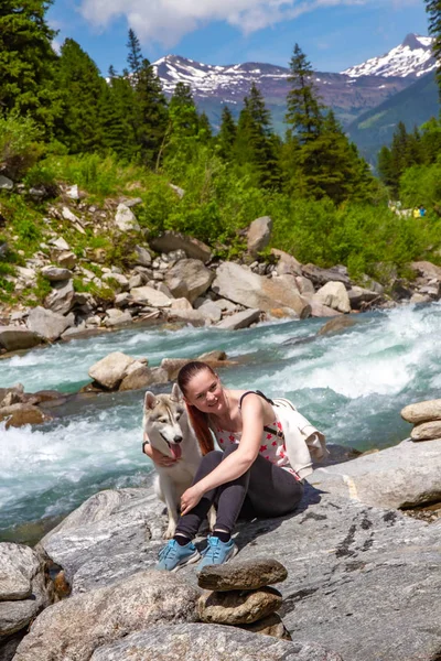 Egy fiatal nő barna hajjal és fehér pulóverrel ül a mólónál a tónál nyugodt vízzel. Egy szibériai husky nőstény kutya fekszik a lány mellett. Az Alpok hegyek a háttérben. — Stock Fotó