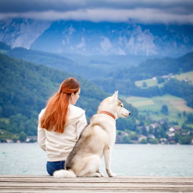 Kahverengi saçlı ve beyaz kazaklı genç bir kadın göl kenarındaki rıhtımda sakin suyla oturuyor. Sibiryalı iri yarı bir dişi köpek kızın yanında uzanıyor. Arkadaki Alpler dağları.