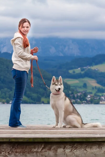 Een jonge vrouw met bruin haar en witte trui zit aan de pier bij het meer met rustig water. Een Siberische husky teef ligt naast het meisje. De Alpen op de achtergrond. Rechtenvrije Stockfoto's