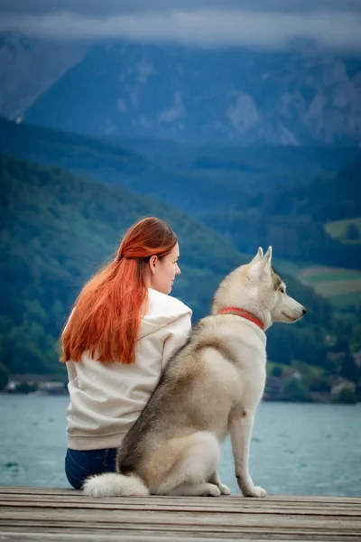 Una joven con cabello castaño y suéter blanco está sentada en el muelle del lago con el agua tranquila. Una perra hembra husky siberiana está acostada cerca de la chica. Las montañas de los Alpes al fondo . —  Fotos de Stock