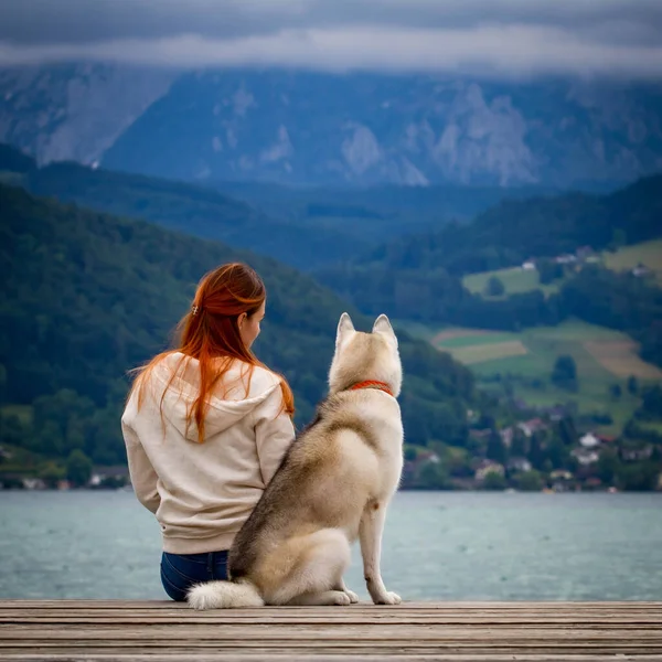 A young woman with brown hair and white sweater is sitting at the pier at the lake with calm water. A Siberian husky female dog is lying down near the girl. The Alps mountains in the background. — Stock Photo, Image