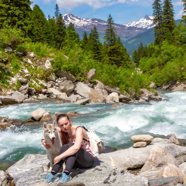 Kahverengi saçlı ve beyaz kazaklı genç bir kadın göl kenarındaki rıhtımda sakin suyla oturuyor. Sibiryalı iri yarı bir dişi köpek kızın yanında uzanıyor. Arkadaki Alpler dağları.