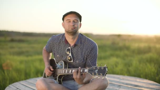 Чоловік грає на електрогітарі в полі на заході сонця — стокове відео