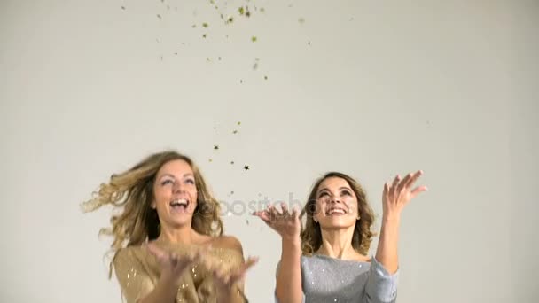 Dos hermosas chicas en blusas brillantes lanzan confeti sobre un fondo blanco — Vídeos de Stock