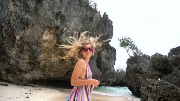 Jong blond meisje in een lichte jurk loopt langs het witte strand, glimlachend in de camera, ontwikkeling van haren. — Stockvideo