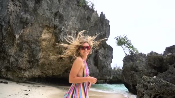 Jovem loira em um vestido brilhante corre ao longo da praia branca, sorrindo para a câmera, desenvolvendo o cabelo . — Vídeo de Stock