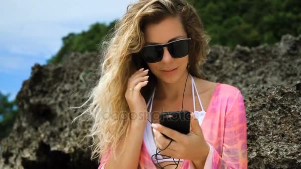 Chica con ropa brillante escuchando música en una playa blanca en piedra. Mujer joven poniendo auriculares en la oreja por la orilla del mar . — Vídeo de stock