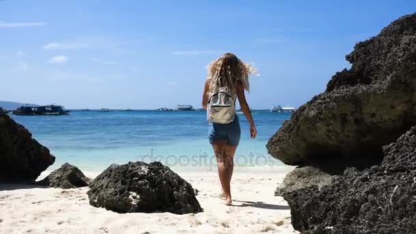 Young girl with beautiful hair in overalls with a backpack goes on a beautiful white beach with rocks — Stock Video