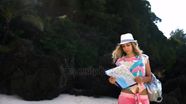 Młoda dziewczyna Blondynka turystycznych w kapeluszu i z plecak patrząc na mapę świata na plaży — Wideo stockowe
