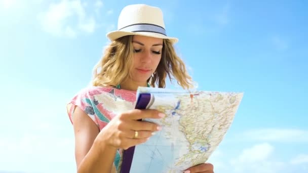 Giovane ragazza turista bionda in cappello e con lo zaino guardando mappa del mondo sulla spiaggia — Video Stock