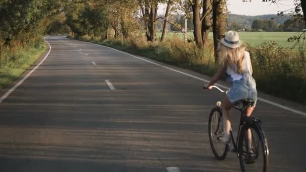 Una giovane ragazza bionda in pantaloni su bici retrò andando lungo la strada nel campo — Video Stock