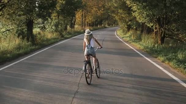 Uma jovem loira de calças na bicicleta retro descendo a estrada no campo — Vídeo de Stock