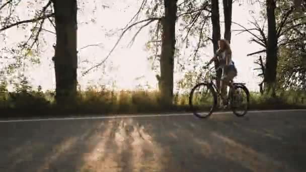 Una giovane ragazza bionda in pantaloni su bici retrò andando lungo la strada nel campo — Video Stock