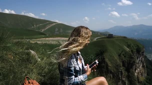 Een jong blond meisje zit op het gras op een achtergrond van bergen met een rugzak en neemt foto's van de bergen op de telefoon — Stockvideo