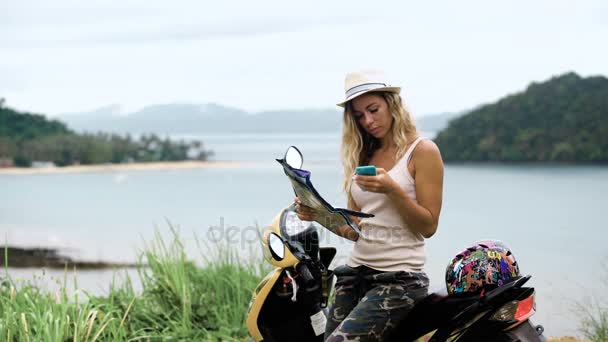 Chica rubia en un sombrero, se sienta en una bicicleta y mira el teléfono y un mapa, mira la ruta en Asia — Vídeos de Stock