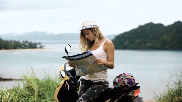 Menina loira de chapéu, senta-se em uma bicicleta e olha para o telefone e um mapa, olha para a rota na Ásia — Vídeo de Stock