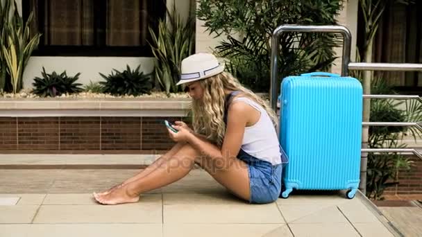 Ξανθιά κοπέλα σε ένα καπέλο, φόρμες με μια μπλε τσάντα, εγκαθίσταται σε ένα τροπικό ξενοδοχείο με πισίνα. 4k — Αρχείο Βίντεο
