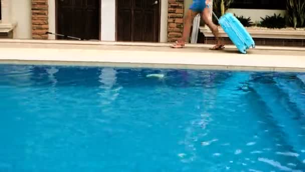 Menina loira em um chapéu, macacão com uma bolsa azul, se instala em um hotel tropical com uma piscina. 4K — Vídeo de Stock
