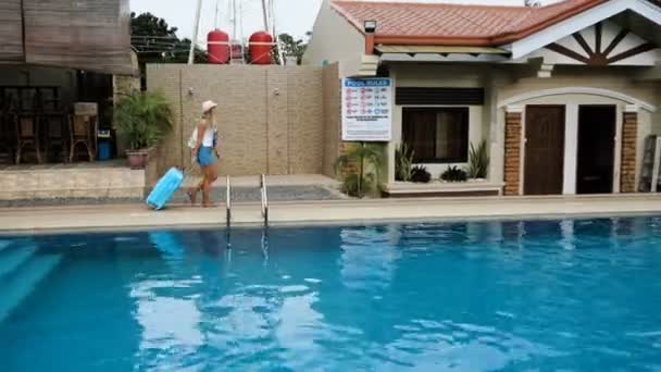 Menina loira em um chapéu, macacão com uma bolsa azul, se instala em um hotel tropical com uma piscina. 4K — Vídeo de Stock