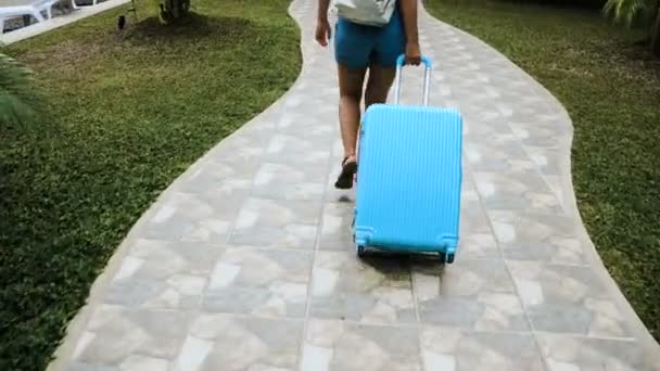 The blond girl in a hat and overalls settles in a tropical hotel with a blue bag. — Stock Video