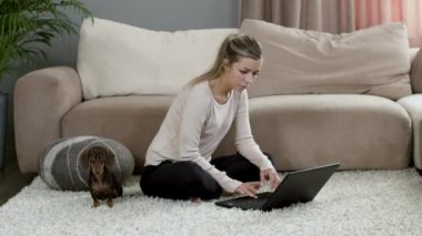 Sarışın kız dizüstü bilgisayarda oturuyor ve evde halıda köpek yavrusu var.