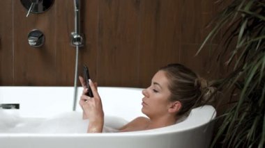 Banyodaki beyaz kız banyoda uzanır ve telefonda selfie çeker..