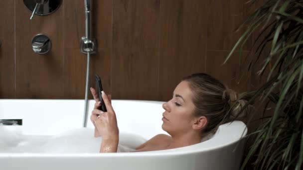 Kaukasische Mädchen im Badezimmer liegt freistehende Badewanne und macht Selfie am Telefon. — Stockvideo