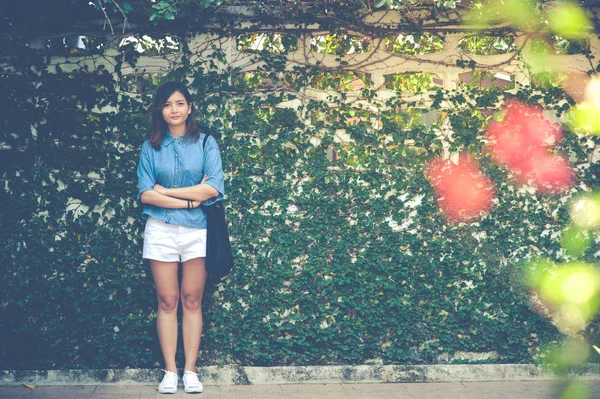 Mujer sonriendo mientras se apoya en la pared al aire libre, feliz mujer elegante hipster de pie en la pared de hojas verdes, concepto de viaje de verano —  Fotos de Stock