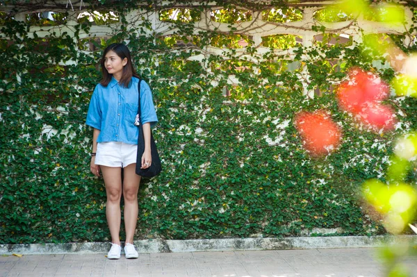 Mujer sonriendo mientras se apoya en la pared al aire libre, feliz mujer elegante hipster de pie en la pared de hojas verdes, concepto de viaje de verano — Foto de Stock
