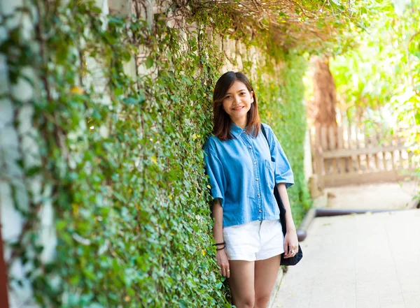 Mujer sonriendo mientras se apoya en la pared al aire libre, feliz mujer elegante hipster de pie en la pared de hojas verdes, concepto de viaje de verano —  Fotos de Stock