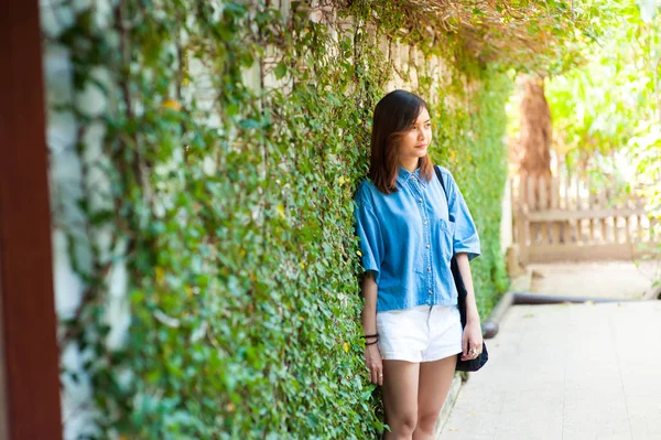 Mujer sonriendo mientras se apoya en la pared al aire libre, feliz mujer elegante hipster de pie en la pared de hojas verdes, concepto de viaje de verano —  Fotos de Stock