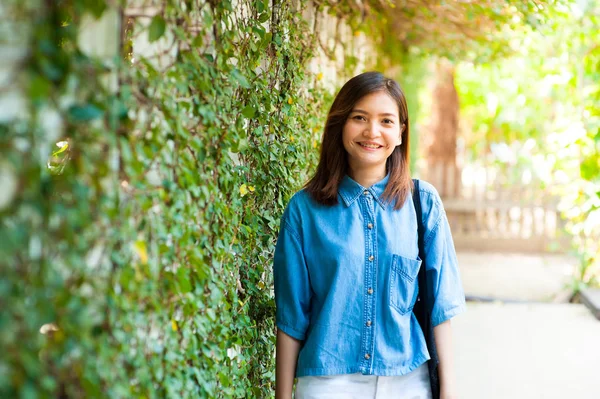 Mujer sonriendo mientras se apoya en la pared al aire libre, feliz mujer elegante hipster de pie en la pared de hojas verdes, concepto de viaje de verano —  Fotos de Stock