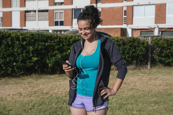 white, caucasian, latin woman, with shorts and sportswear, using her cell phone with headphones, listening music on the grass, before running, turning on a mobile app