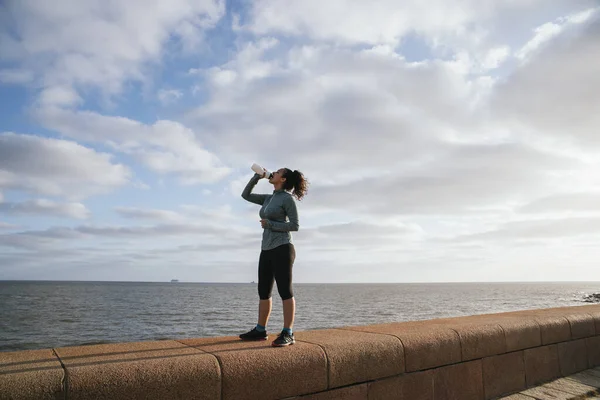 athletic woman drinking water near the sea, with the sky blue with clouds, using sport clothes