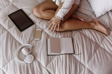 Kız yatakta oturuyor. Tabletteki boşluğu kopyala. Serbest çalışan biri evde çalışıyor. Cep telefonu, not defteri, kulaklık ve tablet yatağın üstünde..