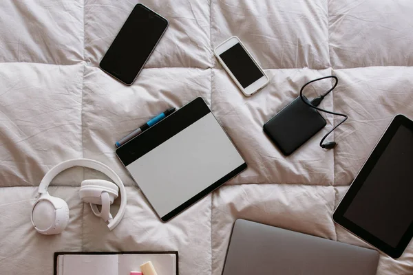 freelance devices on bed. mobile phone, cellphone, external disk, draw tablet, computer, notepad, notebook, headphones, on a white padded.