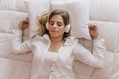 Beyaz, beyaz, beyaz ve sarışın kız, uykulu müzik dinliyor, beyaz kulaklıklı, yastıklı ve beyaz yastıklı, ve bir cep telefonu, bilgisayar, defter.