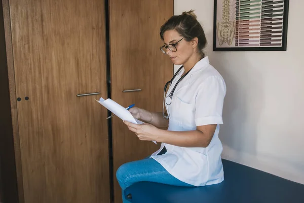 female medical doctor reading papers in her office, with glasses, a stethoscope around her neck and white medical jacket