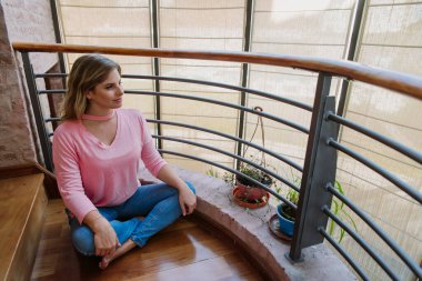 Kafkasyalı, sarışın, genç bir kadın, sakin görünüyor, izliyor, ahşap zeminde oturuyor, nilüfer pozisyonunda, pembe gömlek ve mavi kotla