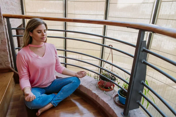 Beyaz, sarışın, beyaz, ahşap zeminde oturan genç kız, nilüfer pozisyonunda, elleri dizlerinde ve gözleri kapalı meditasyon yapıyor, pembe gömlek ve mavi kot pantolon.