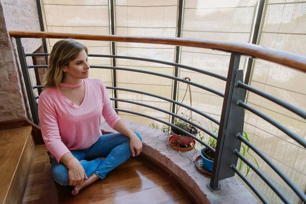 Kafkasyalı, sarışın, genç bir kadın, sakin görünüyor, izliyor, ahşap zeminde oturuyor, nilüfer pozisyonunda, pembe gömlek ve mavi kotla