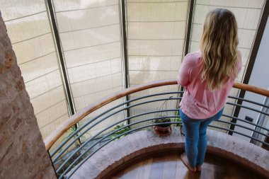 young blond woman from behind, looking at the window in the wooden stair break, barefoot, dressed with pink shirt and blue jeans, rear view from above clipart