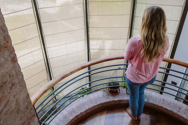 young blond woman from behind, looking at the window in the wooden stair break, barefoot, dressed with pink shirt and blue jeans, rear view from above