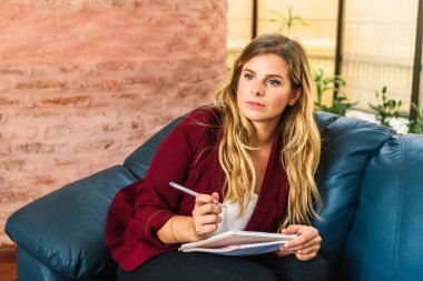 Kafkasyalı genç bir kadının elinde kalemle evdeki mavi bir koltukta otururken çekilmiş resmi..