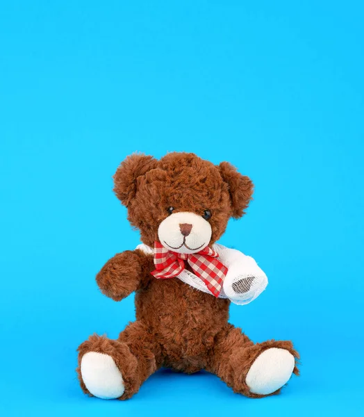 Brown teddy bear with rewound white bandage paw on a blue backgr — Stock Photo, Image