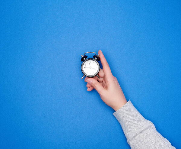 hand with a gray sweater holds a round black alarm clock