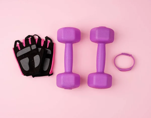 Para fioletowych plastikowych hantli i czarne rękawiczki na różowym grzbiecie — Zdjęcie stockowe